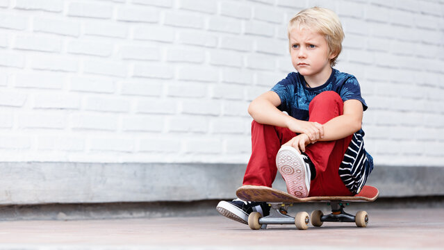 Looking unhappy and upset skater sit on skateboard before children training class in skate park. Active family lifestyle, outdoor recreational activities, kids sports on summer holidays in city.