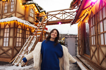 Obraz na płótnie Canvas Friendly young caucaisan girl posing at camera at amusement park with decorate house on background. Brunette wears caual winter clothes. Concept of funny moments, weekends.