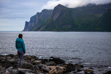 Fototapeta na wymiar girl in blue jacket stands on a rocky beach admiring the mighty mountains on the island of senja, norway; hiking in the norwegian fjords