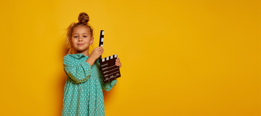 Lovely smiling little girl holding director's film movie slateboard over yellow studio background. Concept of human emotions, beauty, kids fashion, children, school and ad. Copy space.
