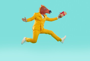 Fototapeta na wymiar Full body side view strange excited man in yellow suit and funny carnival horse mask holding gift present box, hurrying to party, running, jumping on trampoline, flying high on blue studio background