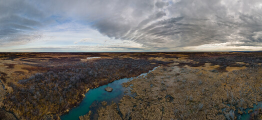 Panorama of a dry landscape of grass and shrubs with a turquoise blue stream under a dramatic sky of grey clouds with patches of blue
