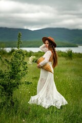 Fototapeta na wymiar A middle-aged woman in a white dress and brown hat stands with her back on a green field and holds a basket in her hands with a large bouquet of daisies. In the background there are mountains and a
