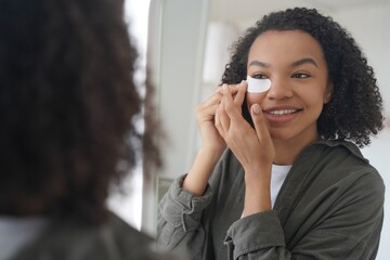 Smiling young mixed ace girl applying under eye patches looking in mirror. Skincare, beauty routine