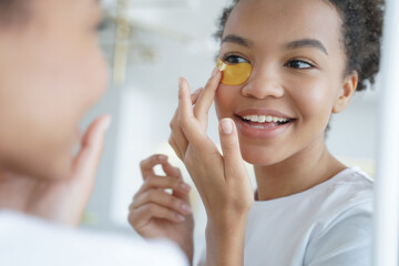 Young mixed race girl applies under eye patches for face skin, looking in mirror. Skincare treatment