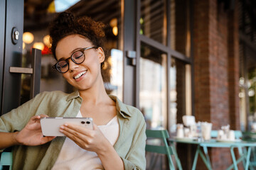 Hispanic woman in eyeglasses smiling and using mobile phone at cafe