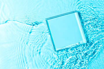 Product presentation background. Mockup glass pedestal or glass stage lying in transparent blue water with splashes. Top view, copy space.