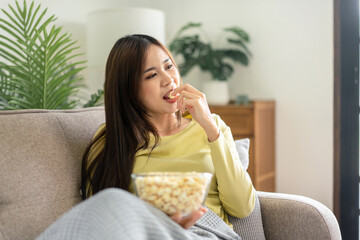 Young asian woman holding a bowl of popcorn to eating popcorn while lying on the big comfortable sofa