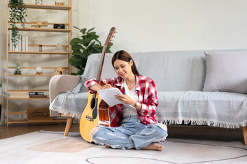 Young woman is compose song and writing lyrics on notebook while putting guitar on the legs and...
