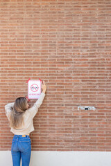 Vertical view of young woman from the back putting a prohibition sign in a brick wall with a drone...