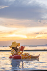 Vertical photo of Happy tourist woman in casual clothes hat, reading a book sitting on red inflatable ring, relaxing by the pool in a luxurious beachfront hotel at sunset enjoying beach vacation.