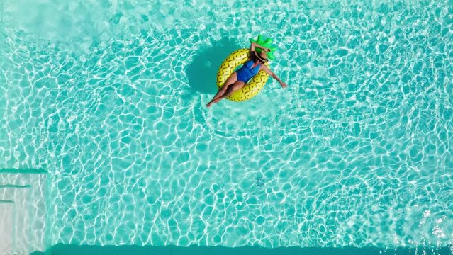 4K drone footage of beautiful woman dressed blue swimsuit floating on yellow inflatable pineapple ring in swimming pool. Resting, summer holidays, free time concept.