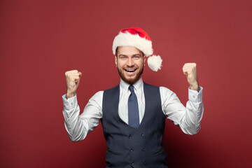 Happy businessman in suit and Christmas hat smiling against red studio wall background. Business...