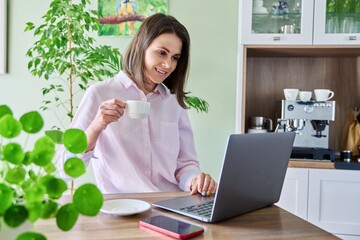 Young woman working at home using laptop, looking at camera, drinking coffee.