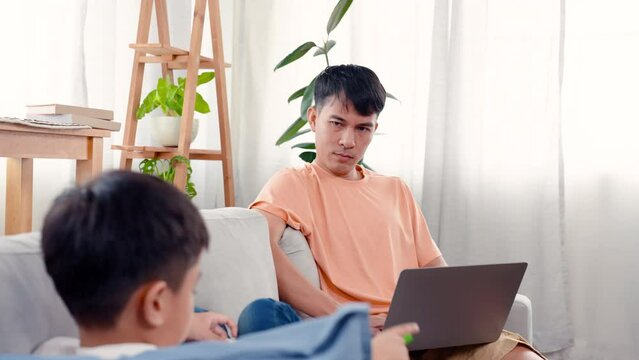 4K, An Asian single father is working on the sofa in the living room of the house and his son is sitting on the other side of the sofa. The father periodically looks at his son.