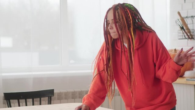 Young woman with bright dreadlocks dancing, wiping table with rag in kitchen. Cheerful female having fun, cleaning at home.