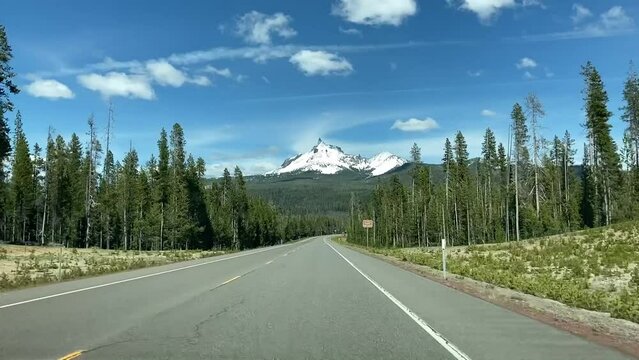 Driving a rural road in Oregon near Crater Lake with a view of snow-covered Mt Thielsen. Driver's point of view through windshield, surrounded by evergreen forest - Oregon, USA