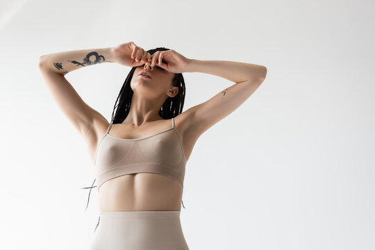 Low angle view of tattooed woman in underwear covering eyes isolated on grey.