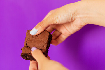 Woman's hand holding a piece of brownie on purple background. Modern food styling composition