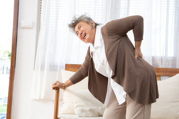 Senior Asian woman is bent over with her hand on the couch because she has lower back pain that is...