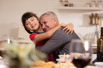 Senior caucasian grandparent hugs his young asian granddaughter while at the dinner table in their home