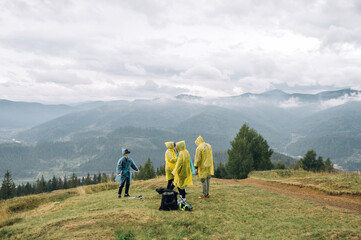 Fototapeta na wymiar A group of young tourists in raincoats are standing in the mountains against the background of a beautiful view during a stopover and are about to continue their journey.