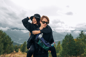 Fototapeta premium A stylish couple of cheerful young tourists are having fun on the top of a mountain, a woman is smiling, a man is carrying her on his back, standing against the background of beautiful mountain views.