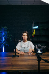 Funny woman sitting at a table and recording videos on camera with a funny face in a professional...