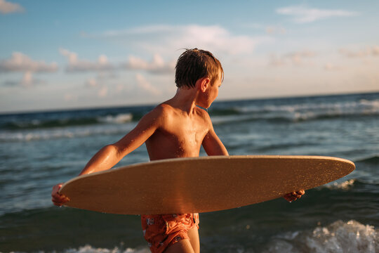 Young boy holding skim board on beach at sunset