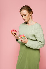 Pensive pregnant woman holding sweet donuts isolated on pink.