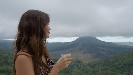 A thoughtful asian girl drinking coffee against fabulous Mountain View with volcano hidden under...