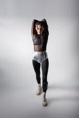 full length of woman in silver shorts over black tight jumpsuit standing with hands behind head on...