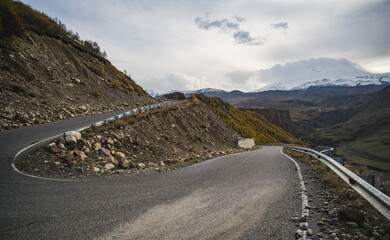 The asphalt serpentine track winds and turns along the mountain slope against the backdrop of a mountain range with snow and glaciers, on a cloudy autumn day in the mountains