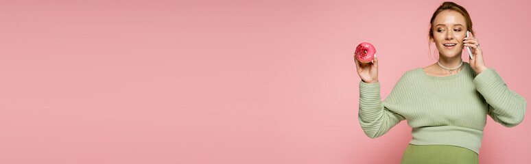Happy pregnant woman talking on smartphone and looking at sweet donut isolated on pink, banner.