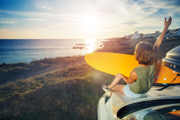 Woman sit on car hood looking at sunset over sea lift up hand