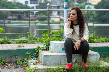 Mid Adult Woman in long hair wearing Sports Clothing sitting outdoors and looking at a distance in garden lake in the background. Front view. Full Length Shot.