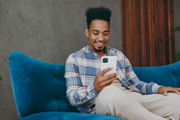 Young man wear casual shirt sits on blue sofa couch hold in hand use mobile cell phone stay at home...