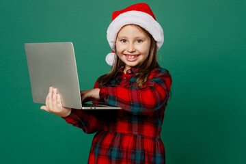 Merry cute little IT child kid girl 6-7 year old wear red dress Christmas hat posing hold use work on laptop pc computer play games isolated on plain dark green background Happy New Year 2023 concept