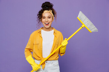 Young fun surprised happy housekeeper woman wear yellow shirt tidy up hold broom sweeps floor clean...