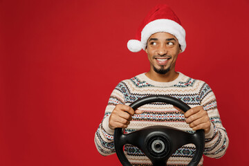 Merry young man wear Christmas sweater Santa hat posing hold steering wheel driving car look aside on workspace area isolated on plain red background. Happy New Year 2023 celebration holiday concept.