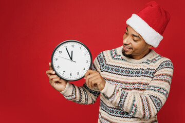 Merry smiling young man wear Christmas sweater Santa hat posing hold show clock time five to twelve waiting midnight isolated on plain red background. Happy New Year 2023 celebration holiday concept.