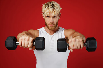 Young strong powerful sporty sportsman man wears white clothes spend time in home gym hold close up dumbbells stretch ahnds to camera isolated on plain red background Workout sport fit body concept.