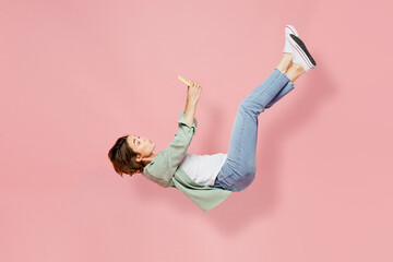 Full body side view young happy fun woman 20s she wear green shirt white t-shirt fly up hover over air fall down use mobile cell phone isolated on plain pastel light pink background studio portrait. - obrazy, fototapety, plakaty