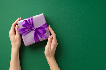Christmas Day concept. First person top view photo of female hands holding lilac giftbox with violet ribbon bow on isolated green background with copyspace