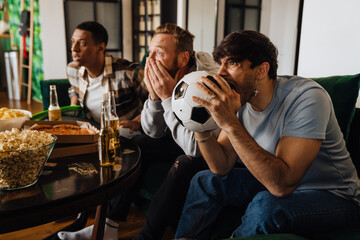 Excited male friends watching sport game and drinking beer while sitting on sofa at home