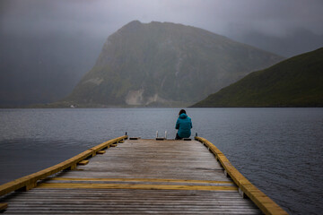 girl sitting at the end of a pier during foggy weather in a Norwegian fjord on lofoten islands;...