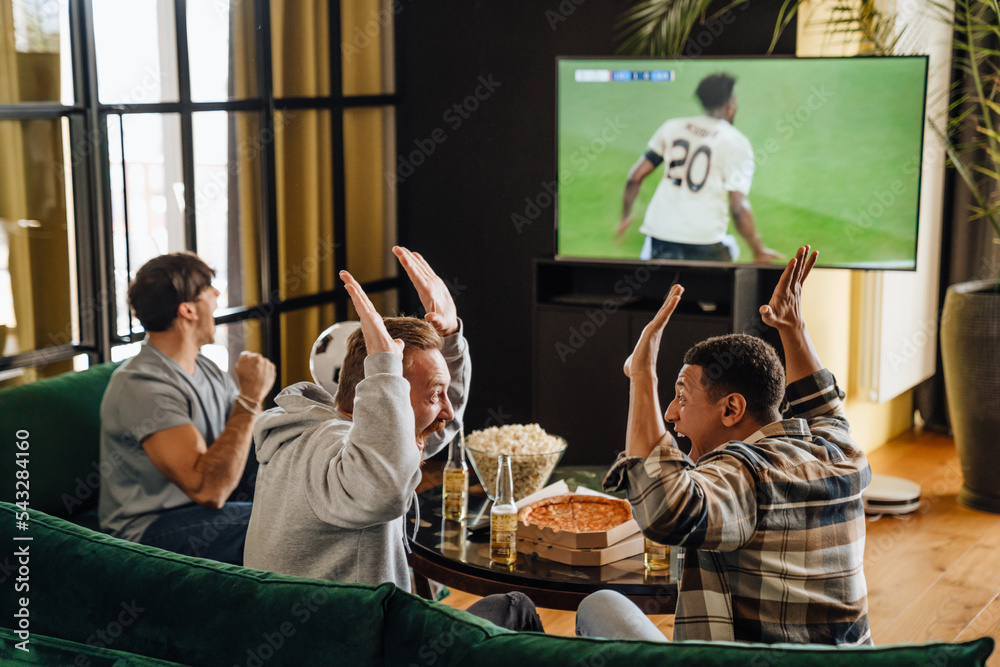 Poster male friends gesturing as winners while watching football match sitting in front of the tv screen