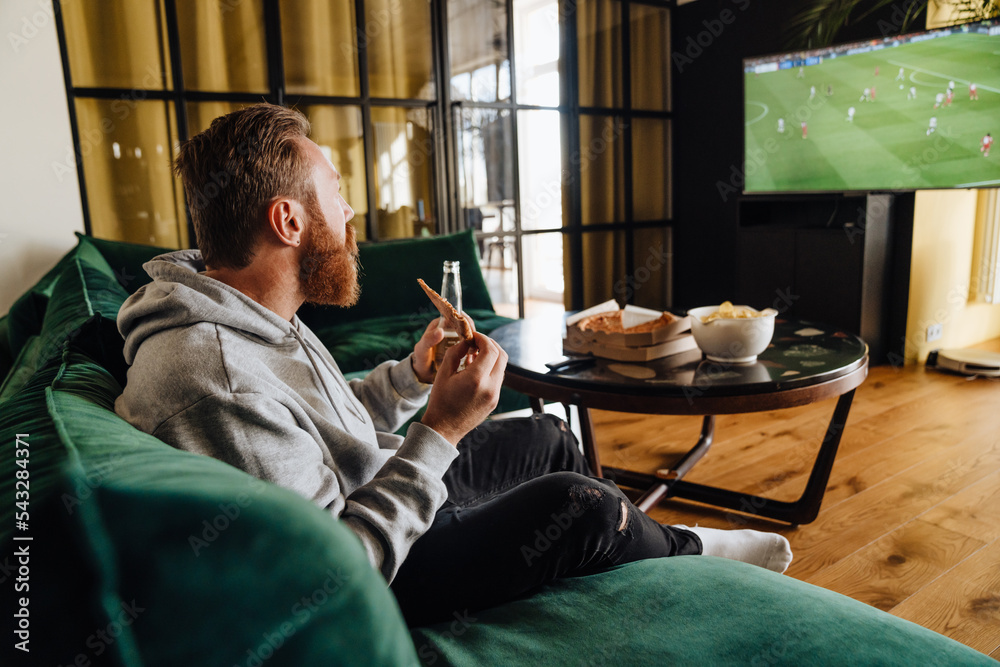 Wall mural Excited white man holding beer and pizza while watching football match