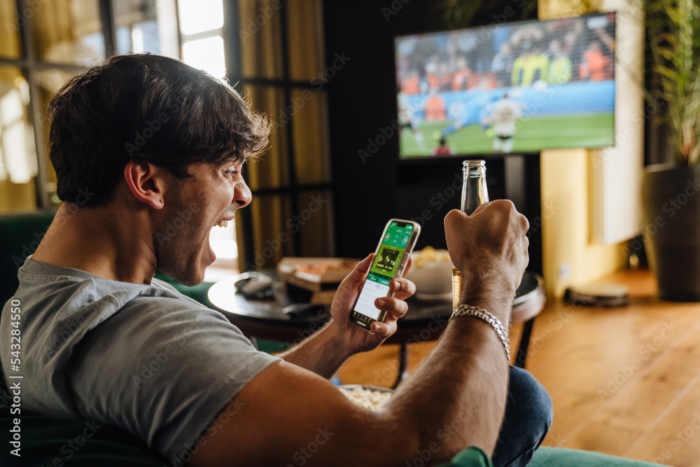 Canvas Prints excited white man watching football match and making bets at bookmaker's website in front of tv scre