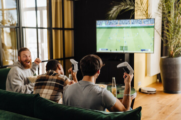 Back view of male friends playing football video game with gamepads at home